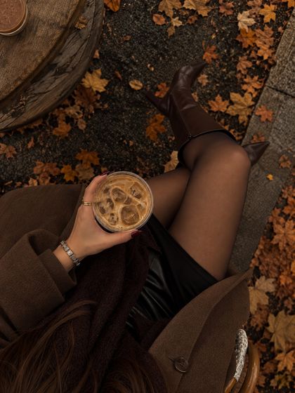 Silver charm bracelet on a wrist holding iced coffee over autumn leaves